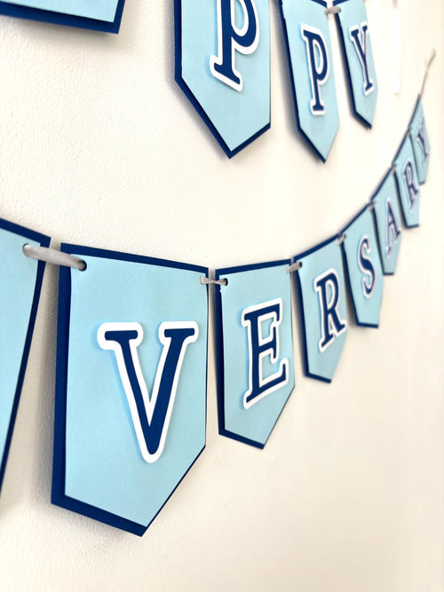 Paper Party Studio Happy Anniversary paper banner with powder blue flags and navy serif letters on a white wall