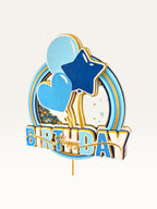 Paper Party Studio blue and gold glitter shaker cake topper with heart, star, and balloon accents.