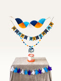 Paper Party Studio blue and yellow birthday decoration set with paper fans, Happy Birthday banner, and cake topper.