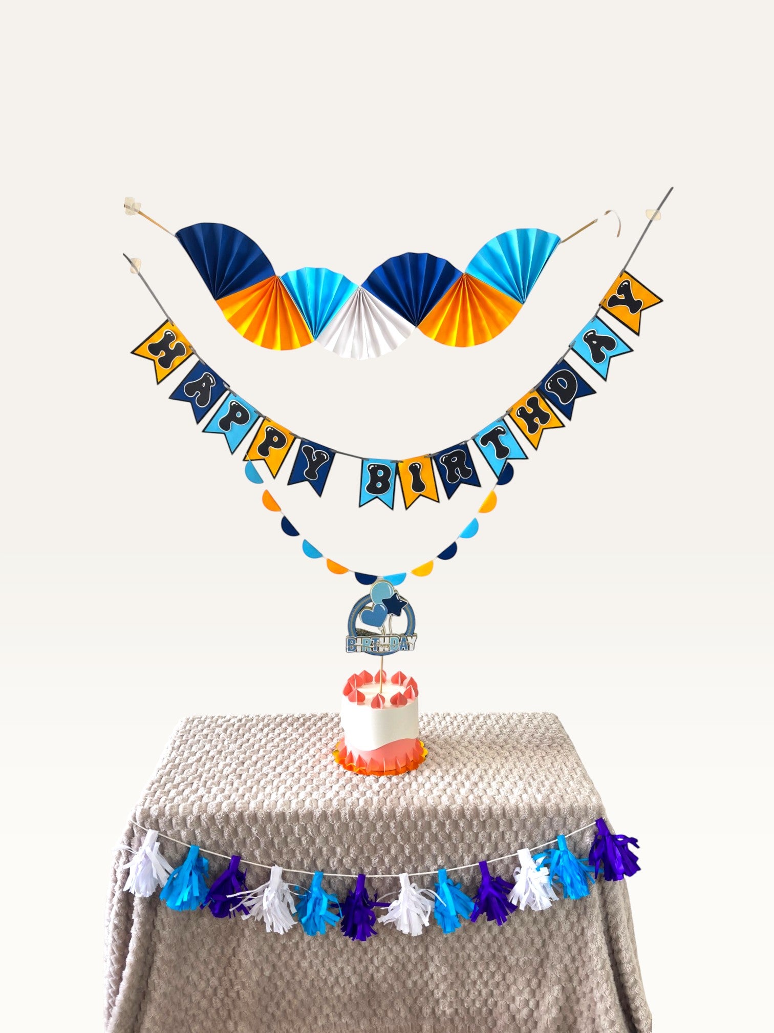 Paper Party Studio blue and yellow birthday decoration set with cake topper, banner, and paper garlands on a table.