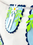 Paper Party Studio Happy Birthday banner with powder-blue scalloped flags, white daisy accents, and green leaves.
