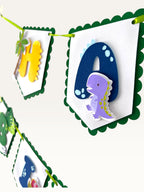 Paper Party Studio dinosaur happy birthday banner with scalloped white flags, green borders, and layered dino letters.