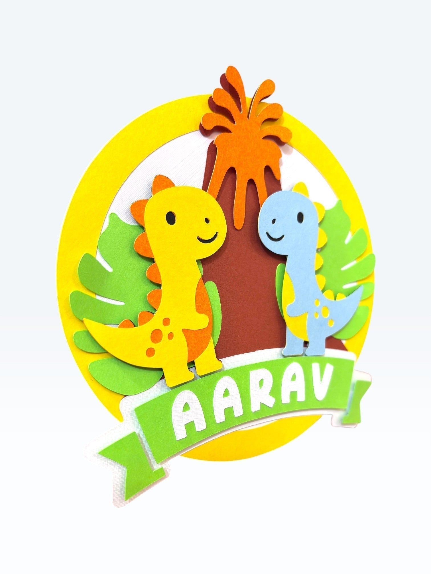 Paper Party Studio personalised dinosaur birthday cake topper with two friendly dinos, volcano, and leaves. Features name 'AARAV'.