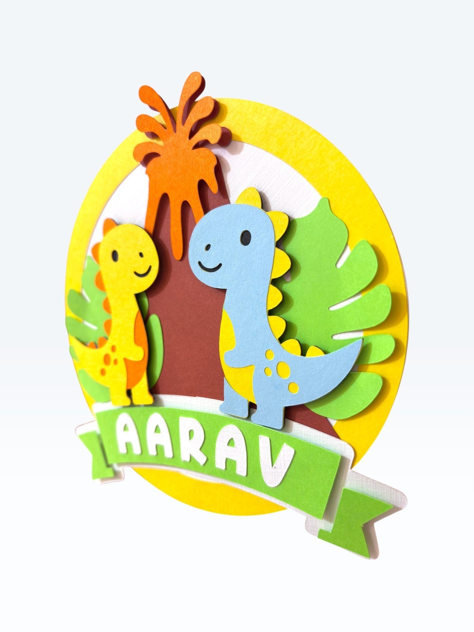 Paper Party Studio personalised dinosaur cake topper with two friendly dinos, volcano, and tropical leaves. Features name 'AARAV'.