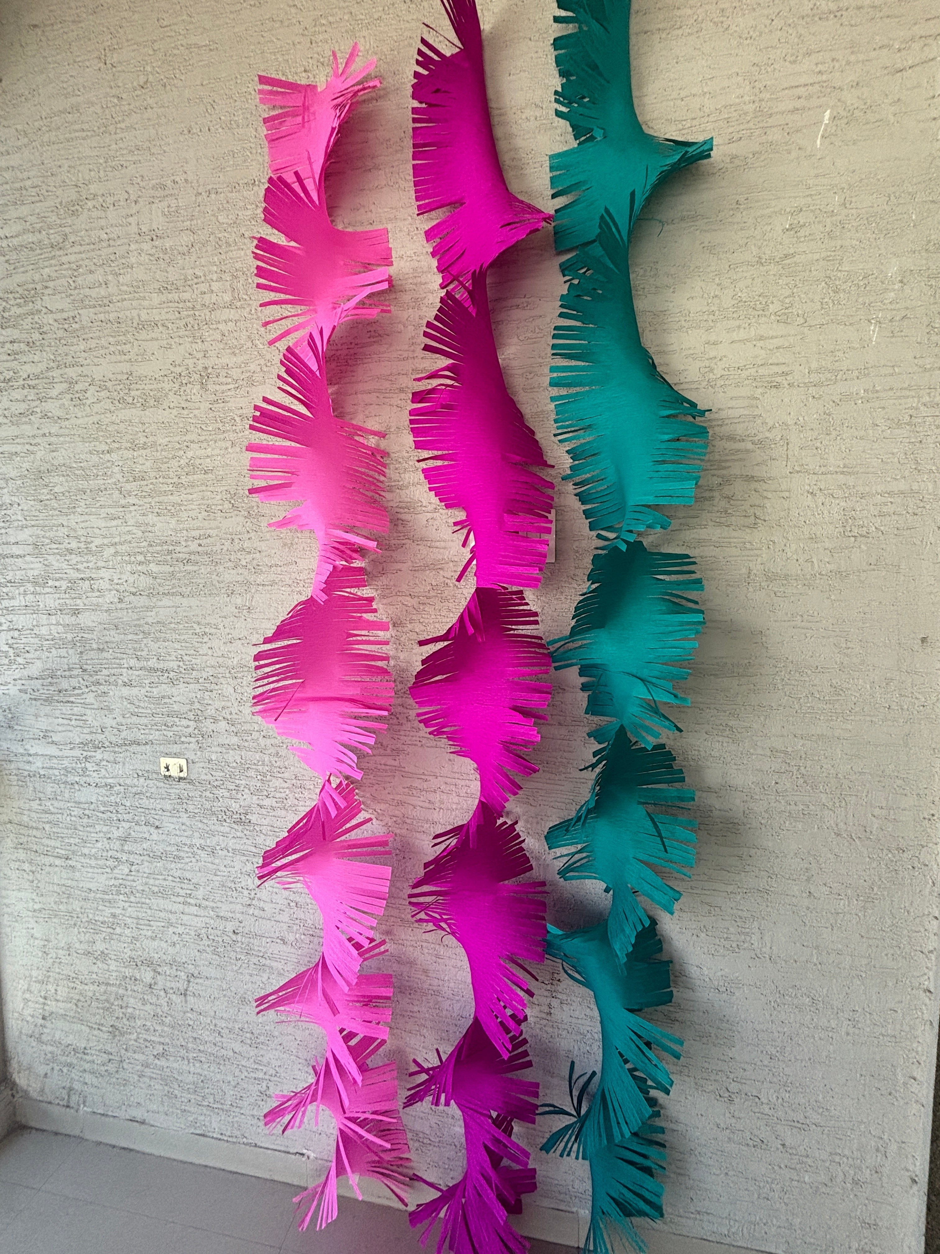 Paper Party Studio pink and teal paper fringe streamers hanging vertically against a textured wall.