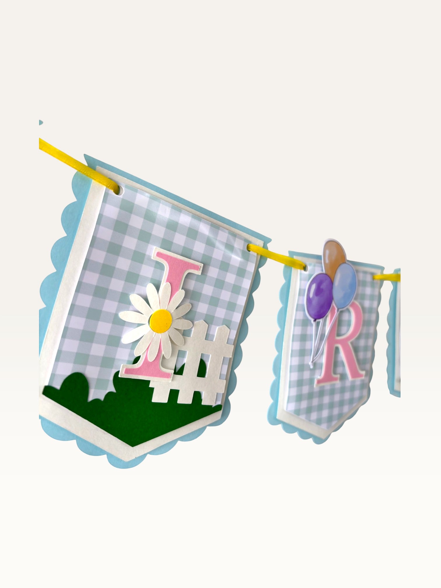 Paper Party Studio gingham garden birthday banner with pastel blue flags, pink letters, daisies, and balloons.