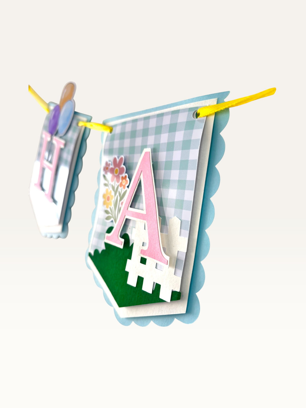Paper Party Studio gingham birthday banner with pastel blue check flags, pink letters, and floral garden decorations.