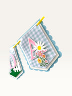Paper Party Studio birthday banner featuring blue gingham paper flags with pink letters, daisies, and wildflowers.