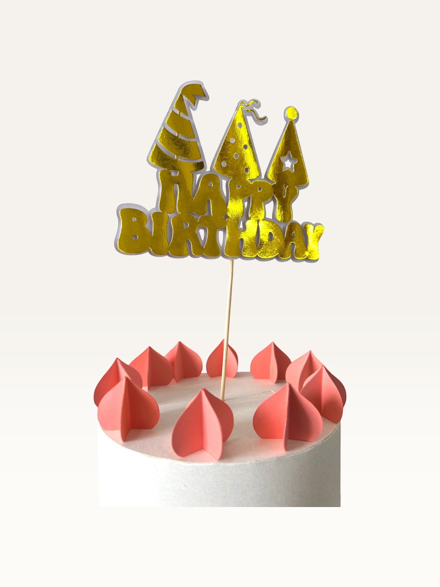 Paper Party Studio golden Happy Birthday cake topper with party hat cut-outs on a white cake with pink decorations