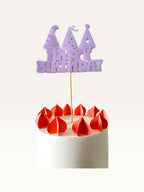Paper Party Studio lavender Happy Birthday cake topper with party hat cut-outs on a wooden pick atop a white cake.