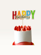 Paper Party Studio multicolour block letter Happy Birthday cake topper with gold script on a white frosted cake