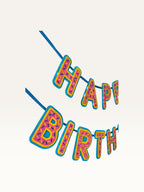 Paper Party Studio Happy Birthday banner with pink donut-glaze letters, candy sprinkles, and yellow outlines.