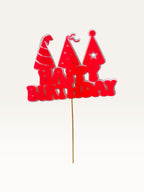 Paper Party Studio red Happy Birthday cake topper with three party hats on a wooden pick against a white background.