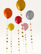 Paper Party Studio set of 10 gold paper star garland strings hanging as decorative tails from colorful balloons.