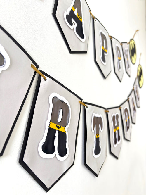 Paper Party Studio superhero themed Happy Birthday paper banner with charcoal and gold letters on silver pennants.