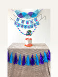 Paper Party Studio tropical luau birthday set with blue alien cake topper, paper fans, banner, and tassel garland.