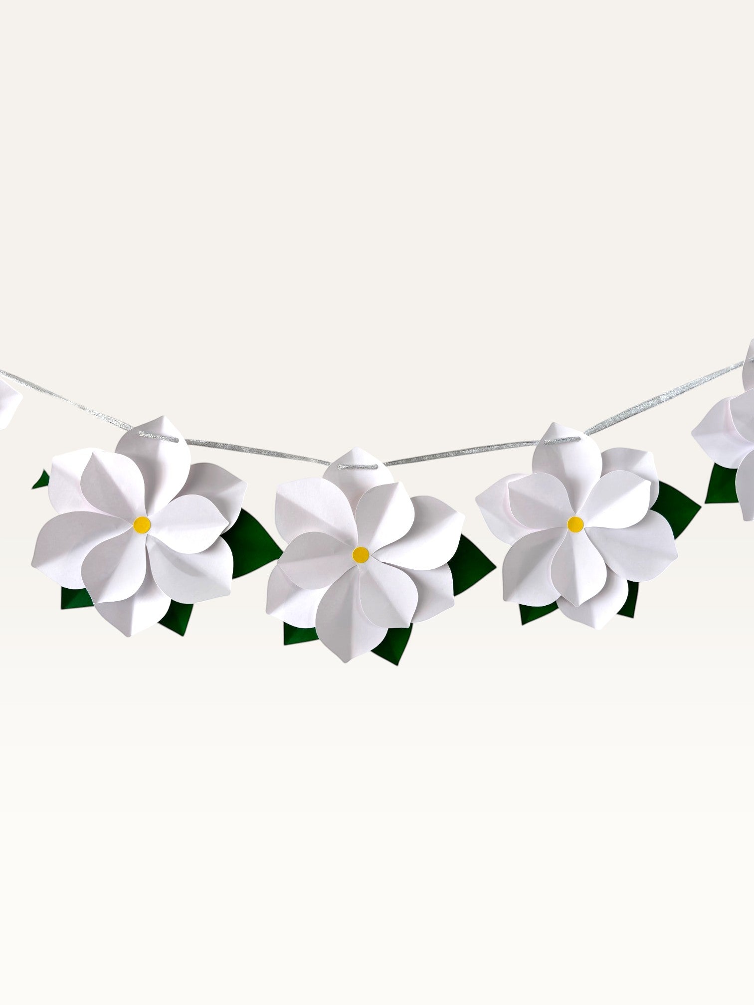 Paper Party Studio white 3D paper flower garland with yellow centers and green leaves on a silver cord for decor