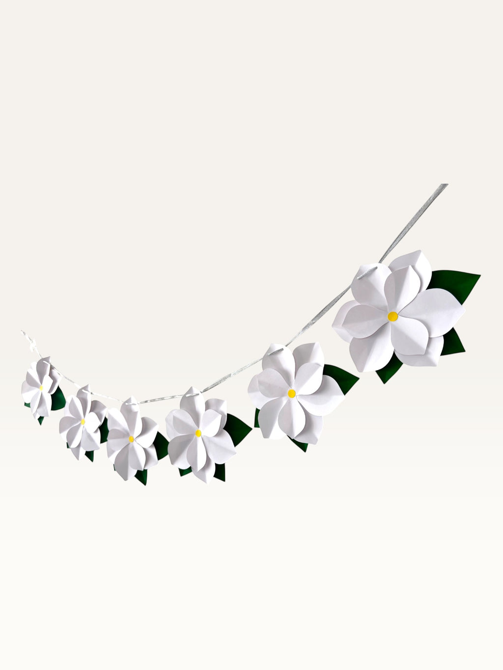 Paper Party Studio white paper flower garland with yellow centers and green leaves on a silver cord for wall decor.