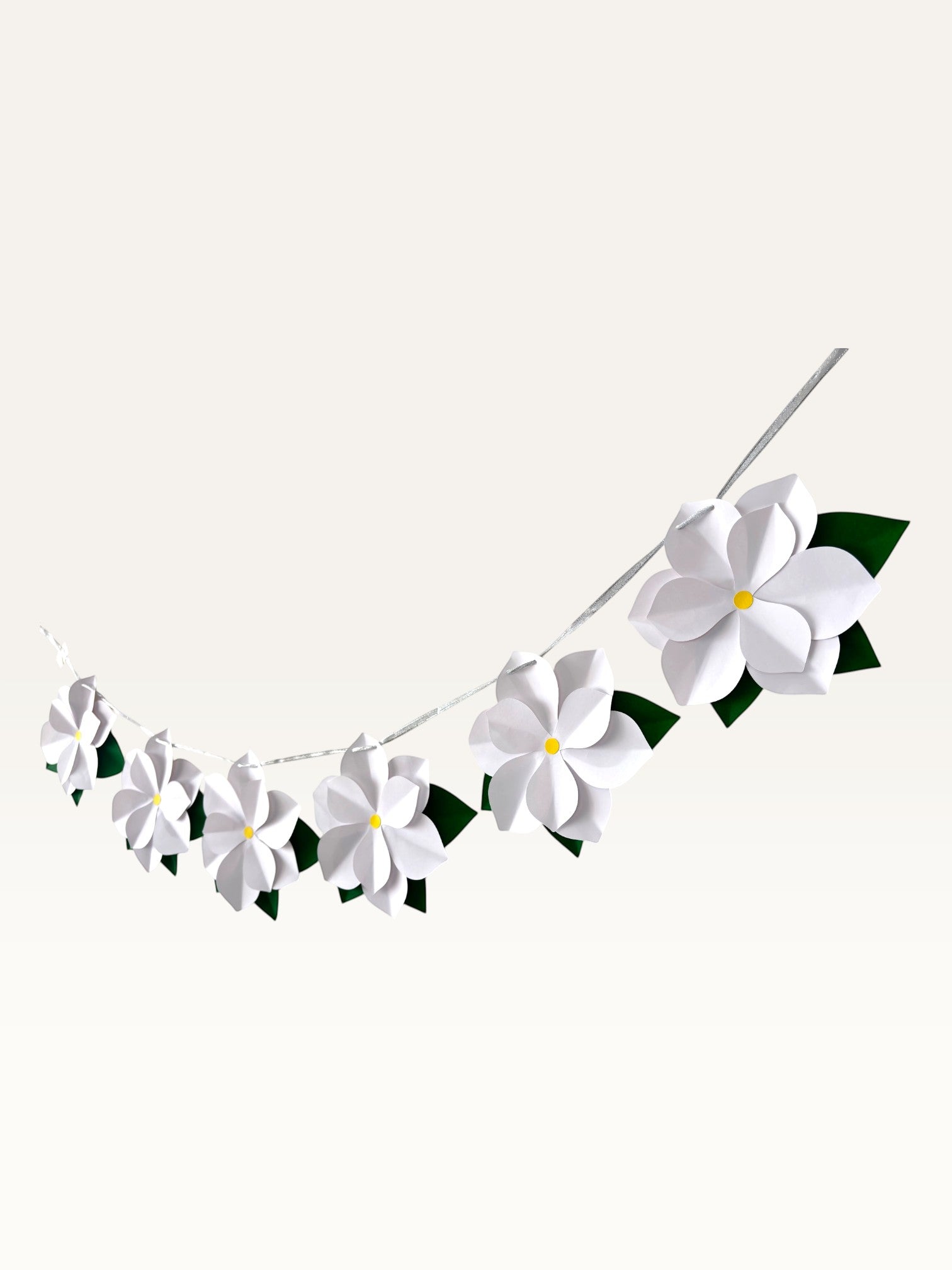 Paper Party Studio white paper flower garland with yellow centers and green leaves on a silver cord for wall decor.